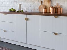 a kitchen with white cabinets and wooden counter tops next to a brick wall, has copper handles on the doors