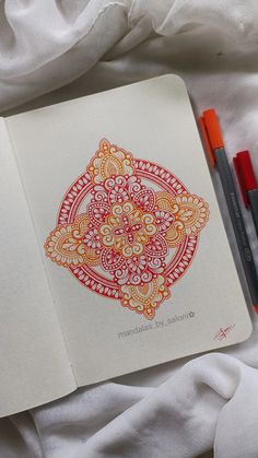 an open notebook with red and orange designs on it next to two markers in front of white sheets