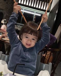 a little boy holding two wooden sticks above his head while sitting in a high chair