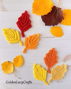 crocheted autumn leaves are arranged on a white wooden surface with yellow, red and orange leaves