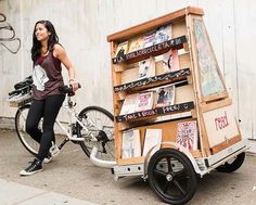 a woman standing next to a cart with books on it and a bicycle parked in front of it