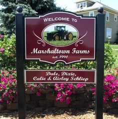 a welcome sign for a farm with pink flowers in the foreground and a large house behind it