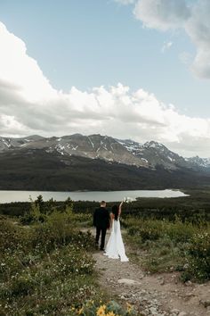 a bride and groom walking down a path towards a lake with mountains in the background