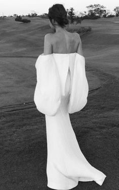 a woman in a long white dress is walking on the grass with her back to the camera