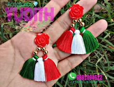 red, white and green tasseled earrings with roses on the end are held in someone's hand