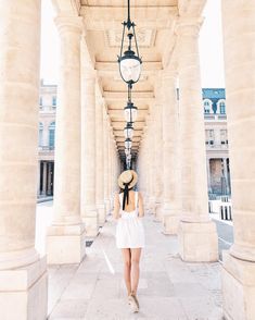 a woman in a white dress and straw hat walking down an empty walkway with columns