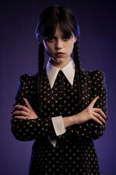 a woman with her arms crossed wearing a black dress and white collared shirt, standing in front of a purple background