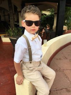 a young boy wearing sunglasses and suspenders sitting on a ledge