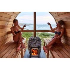 two women in bathing suits sitting on wooden benches near an open fire place with a view of the ocean