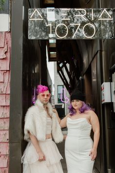 two people standing in front of a building wearing white dresses and fur stoles with pink hair