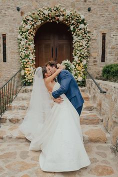 a bride and groom kissing in front of a stone building with flowers on the wall