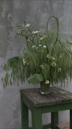 a green table topped with a vase filled with flowers next to a gray wall and wooden bench