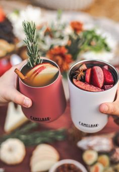 two people holding mugs filled with food on top of a table covered in fruit