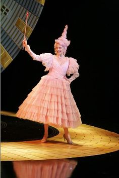 a woman in a pink dress and hat on stage holding a stick with her right hand