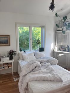 an unmade bed with white sheets and pillows in a bedroom next to two windows