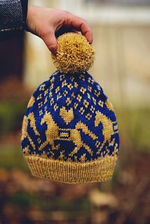 a hand holding a blue and yellow knitted beanie with an animal on it