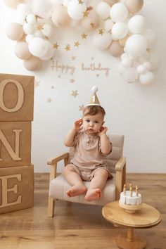 a baby sitting in a chair with a birthday hat on and balloons behind it that spell out happy birthday