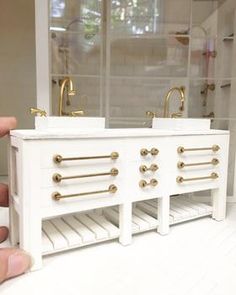 a hand holding a miniature white bathroom vanity with two sinks and gold faucets