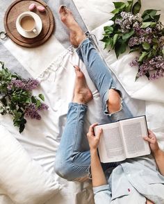 a person laying in bed with an open book next to flowers and a cup of coffee