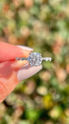 a woman's hand holding an engagement ring with a diamond on it, in front of some bushes