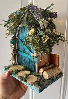 a hand holding up a miniature house made out of wood and plants on top of it