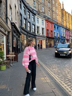 a woman in pink sweater and black pants standing on street next to building with cars