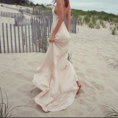 a woman in a white dress is standing on the sand near a fence and grass