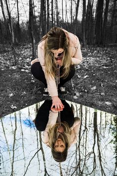 a woman kneeling down in front of a puddle with her reflection on the ground and trees behind her