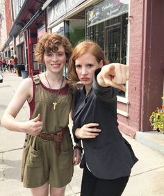 two women are posing for a photo on the sidewalk with one pointing at the camera