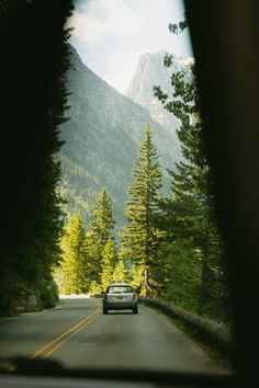 a car is driving down the road in front of some tall trees and mountain peaks