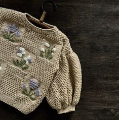 a sweater with flowers is hanging on a hanger against a wooden background that looks like it has been hand knitted