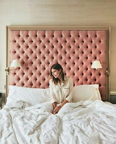 a woman sitting on top of a bed in front of a pink headboard with white sheets