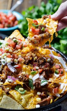 a hand holding a tortilla chip over a bowl of nachos