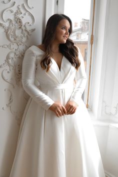 a woman standing in front of a window wearing a white dress with long sleeves and a bow at the waist