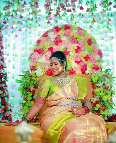 a woman sitting on top of a bed wearing a yellow and green sari with flowers behind her