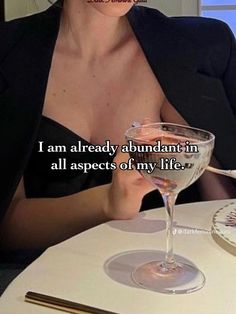 a woman sitting at a table with a wine glass in front of her and the words i am already abundant in all aspects of my life