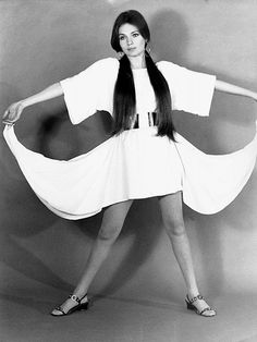 a woman with long hair in a white dress and sandals posing for the camera while holding her arms out