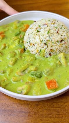 a white bowl filled with green soup and rice on top of a wooden table next to a person's hand