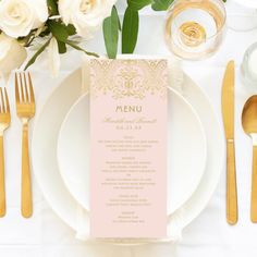 a table setting with pink menu cards and gold cutlery, white flowers and greenery