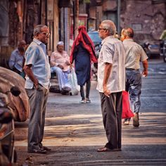 two men are standing on the street talking to each other while others walk down the sidewalk