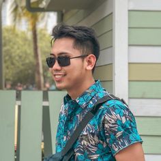 a man with sunglasses and a blue shirt is holding a black bag in front of a house