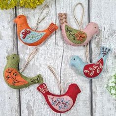 four felt bird ornaments hanging from clothes pins on a white wooden surface with flowers in the background