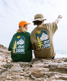 two people sitting on rocks near the ocean pointing at something in the sky with their hands