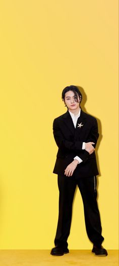 a man in a black suit and white shirt standing against a yellow wall with his arms crossed