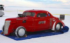 an old red car is parked in the snow on a blue tarp with other cars behind it