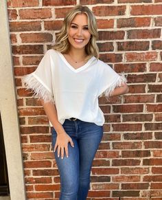 a woman standing in front of a brick wall wearing jeans and a white top with fringe sleeves