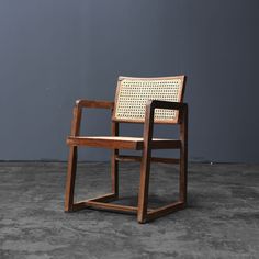 a wooden chair sitting on top of a cement floor next to a gray wall with a woven seat