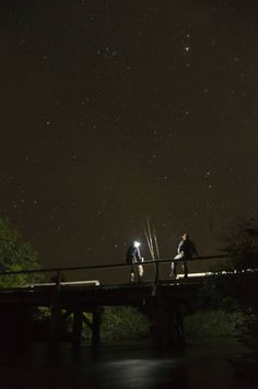 two people standing on a bridge looking up at the sky with stars in the background