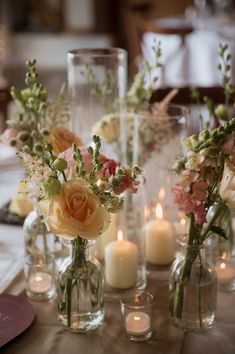 Wedding table centre of pretty peach and pink flowers in bud vases with candles in glass vases. Candlesticks And Bud Vases Wedding, Single Rose Centerpiece With Candles, Bud Vase Centerpiece With Candles, Single Roses In Vases Wedding, Bud Vases With Pampas, Sweetheart Table Bud Vases And Candles, Tealight Wedding Centerpiece, Bud Flowers Wedding Table, Round Wedding Table Bud Vases