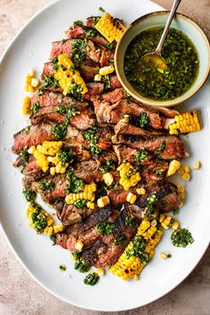 a white plate topped with steak and corn next to a bowl of pesto sauce